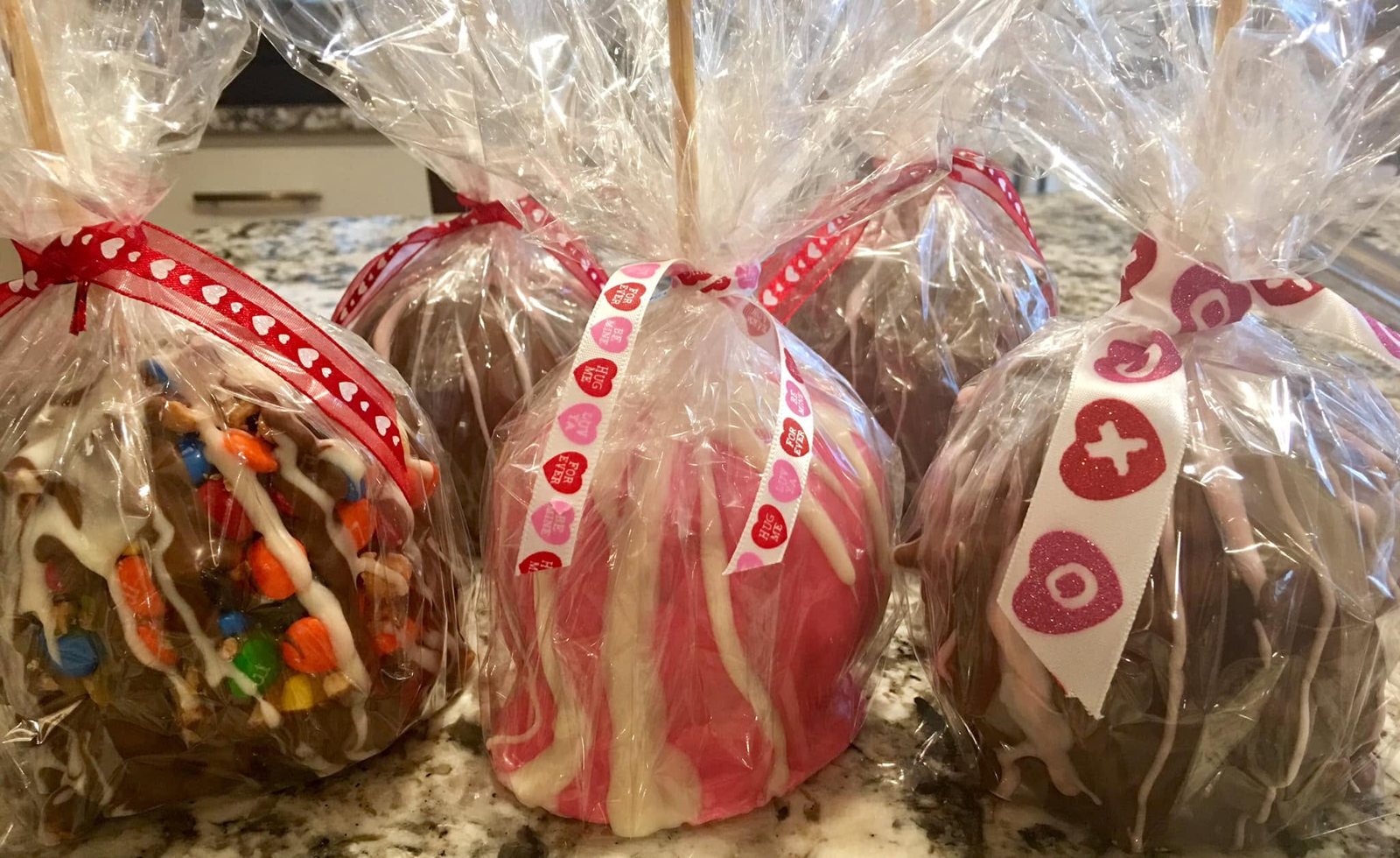 Valentine's Day candy apples in cellophane with heart ribbons, including a pink apple with white drizzle and an M&M apple